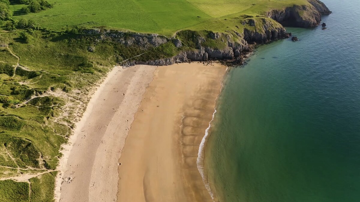 Aerial of Barafundle Bay in Pembrokeshire, a secluded arc of golden sand framed by green headlands and dunes above clear turquoise water