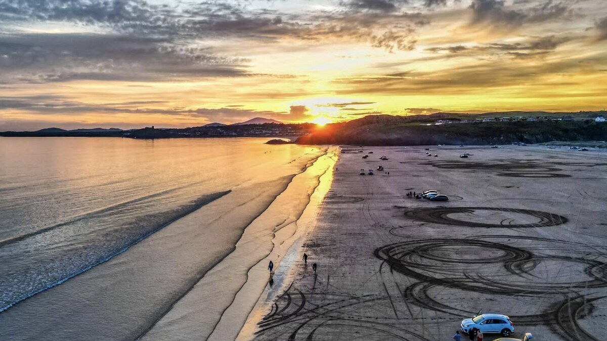 Black Rock Sands near Porthmadog at sunset, cars parked on the firm sand with tyre tracks across it, the sea and hills under a golden sky