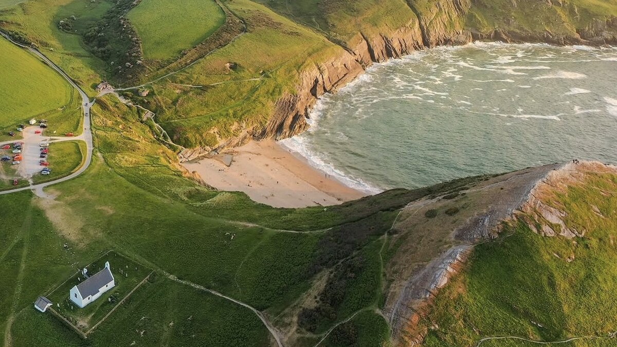 Aerial of Mwnt in Cardigan Bay, a small sandy cove tucked below green cliffs and fields, with the tiny whitewashed church and car park above it