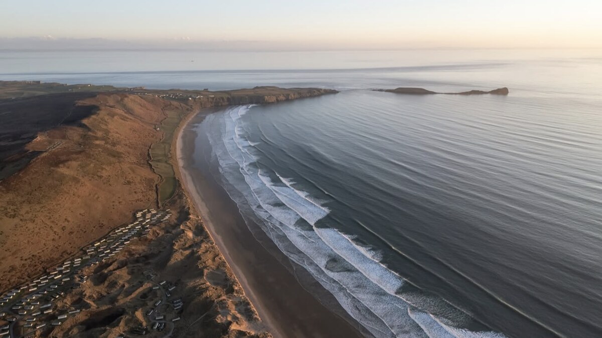 Aerial of Rhossili Bay on the Gower at dusk, the long sweep of sand and rolling surf below the down, with a clifftop caravan park and a headland beyond