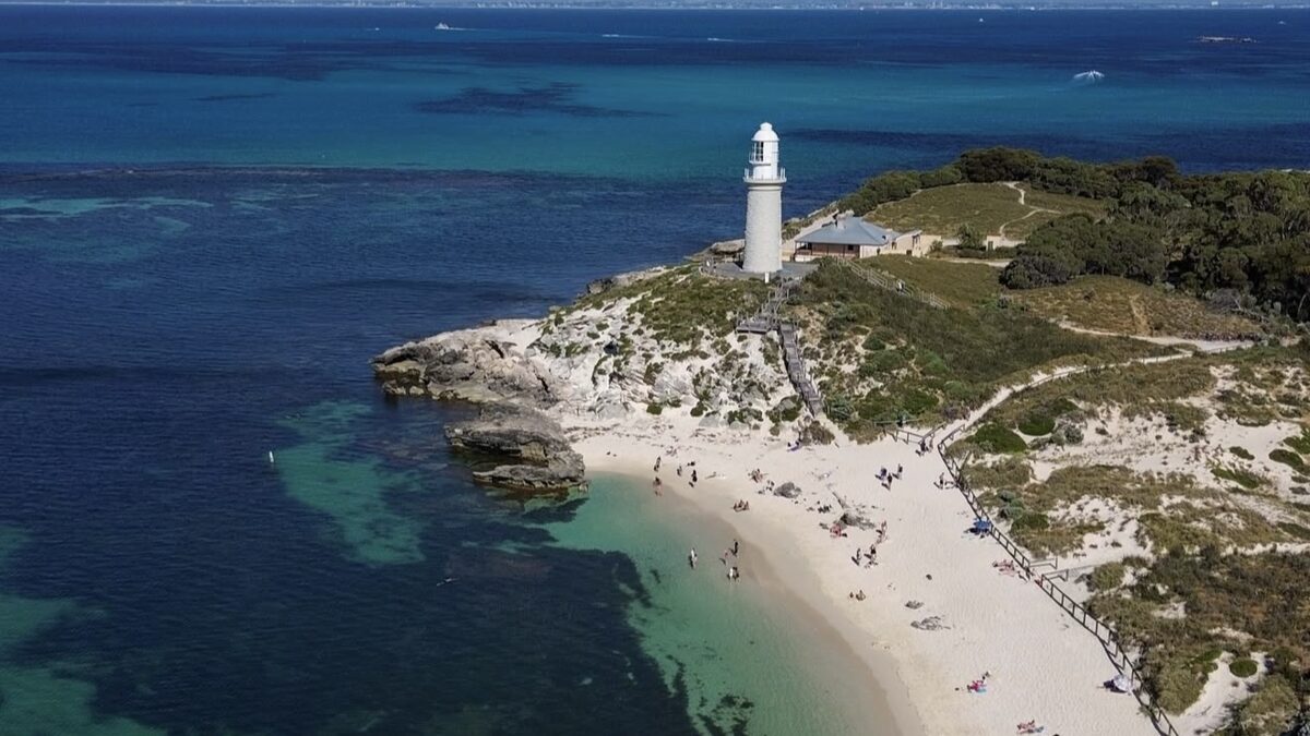 Bathurst Lighthouse above the white sand of Pinky Beach on car-free Rottnest Island, with clear water and the Perth skyline faint on the horizon