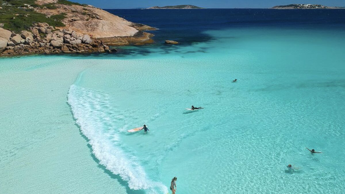 Swimmers in the glass-clear turquoise shallows at Wharton Beach, a granite headland and offshore islands beyond, Duke of Orleans Bay near Esperance