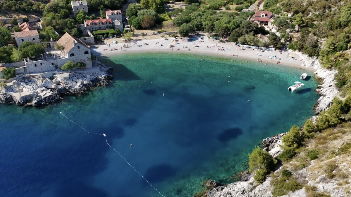 Dubovica beach on Hvar, a small pebble bay of clear turquoise water below a bare hillside, with old stone houses at the water's edge
