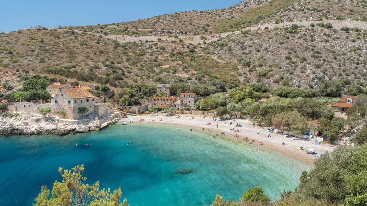 Pupnatska Luka on Korcula, a pebble cove of clear turquoise water backed by old stone houses and bare hills on the island's south coast