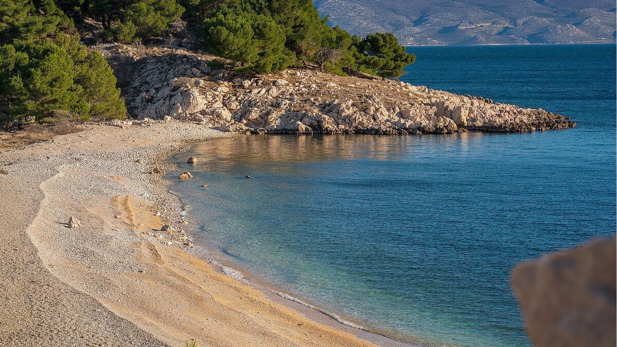 A pebble beach near Baska on the island of Krk, clear water backed by pine-topped rock and the bare Velebit mountains across the channel