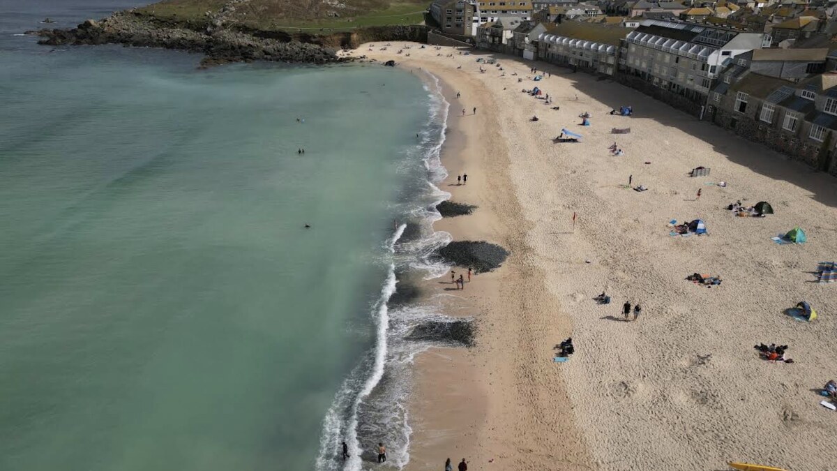 Porthmeor Beach at St Ives from above, surf washing the sand between the town's rooftops and the green headland of the Island