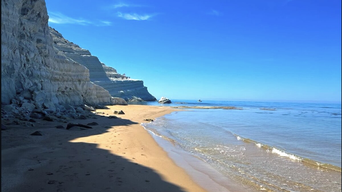 Looking up at the white marl cliffs of Scala dei Turchi from the free beach beside them, the sand in late-afternoon shadow with the calm sea to the right