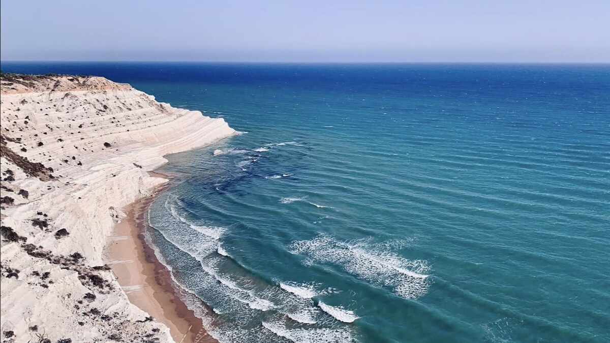 Elevated view along the coast at Scala dei Turchi, the white marl cliffs sloping down to a sandy beach with surf and the deep blue sea stretching along the shore