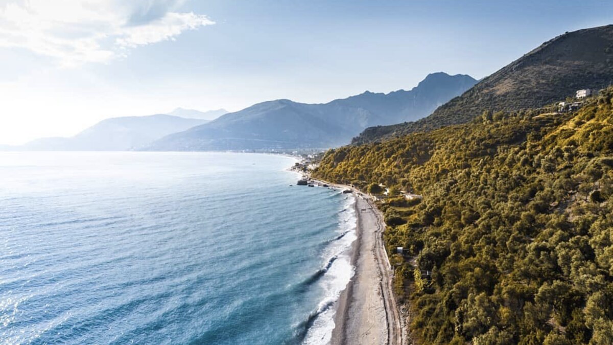 The empty shingle shore at Lukove on the Albanian Riviera, running below olive-covered slopes with the coastal mountains beyond