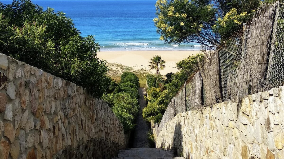 The stone-walled staircase down to Cala de los Alemanes at Atlanterra, with the sand and blue Atlantic framed at the bottom