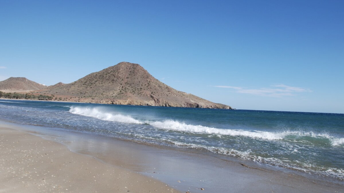 Playa de los Genoveses at beach level, a wave rolling onto empty sand with the rounded Morron headland behind