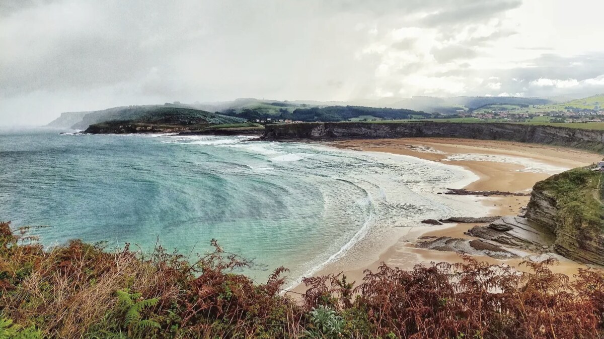 Playa de Langre from the clifftop, golden sand enclosed by dark cliffs and green meadows under a moody Cantabrian sky